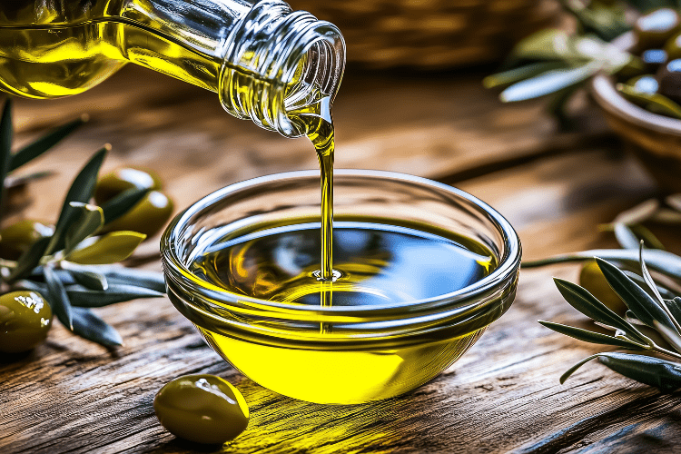 Golden olive oil being poured into a glass bowl on a wooden table.