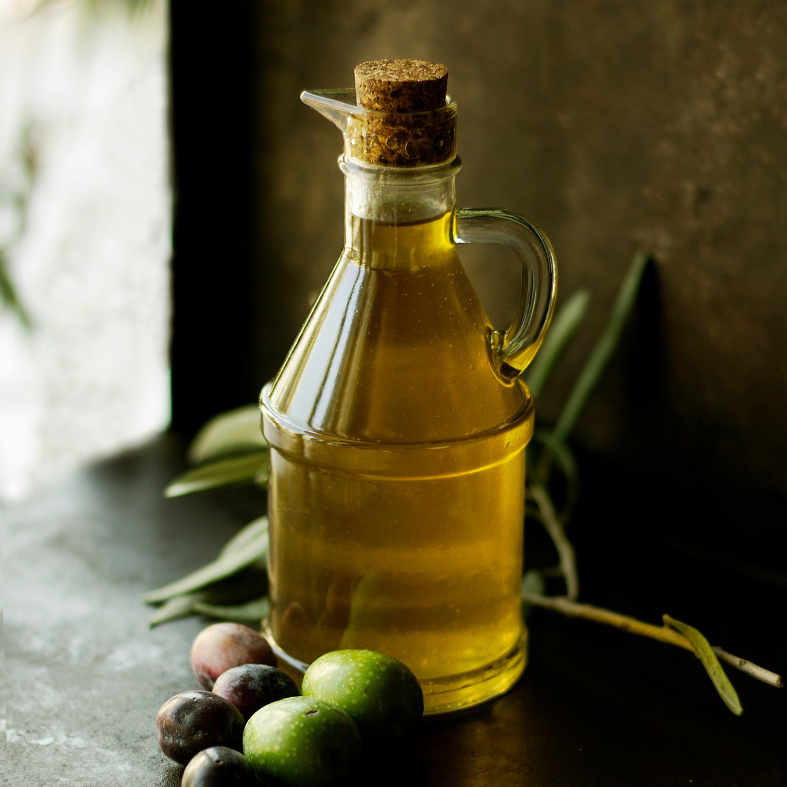 A glass bottle of olive oil with olives and leaves beside it.